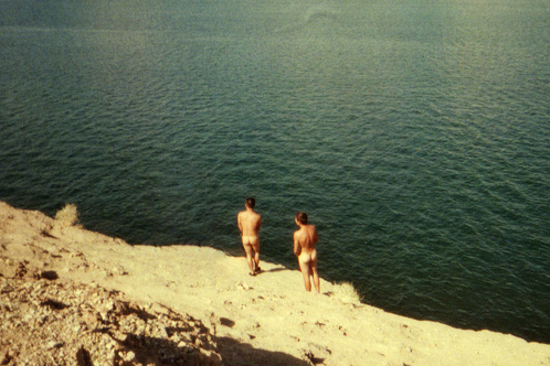 two naked people preparing to recreationally jump from a cliff into a lake