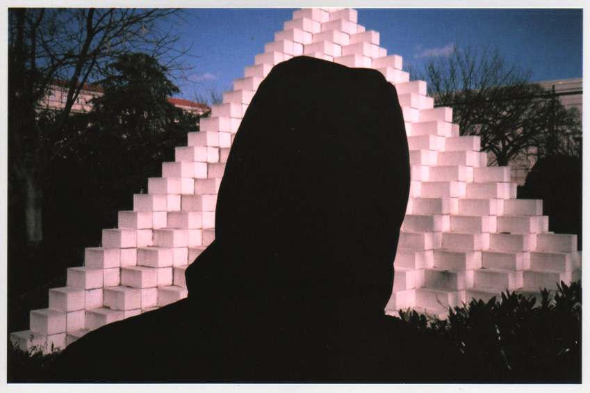 back of hooded figure's head in front of sol lewitt pyramid sculpture